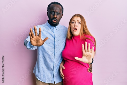 Young interracial couple expecting a baby, touching pregnant belly doing stop gesture with hands palms, angry and frustration expression