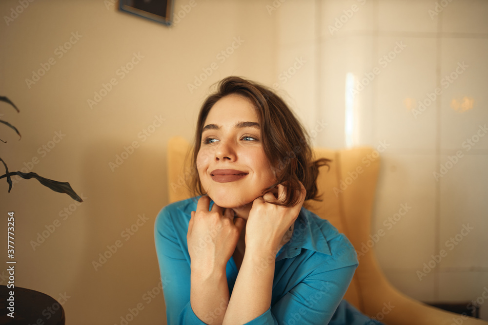 Joy, happiness and relaxation concept. Portrait of gorgeous positive young female in blue dress sitting comfortably in armchair with hands at her face, having dreamy look, thinking about vacations Joy, happiness and relaxation concept. Portrait of gorgeous positive young female in blue dress sitting comfortably in armchair with hands at her face, having dreamy look, thinking about vacations
