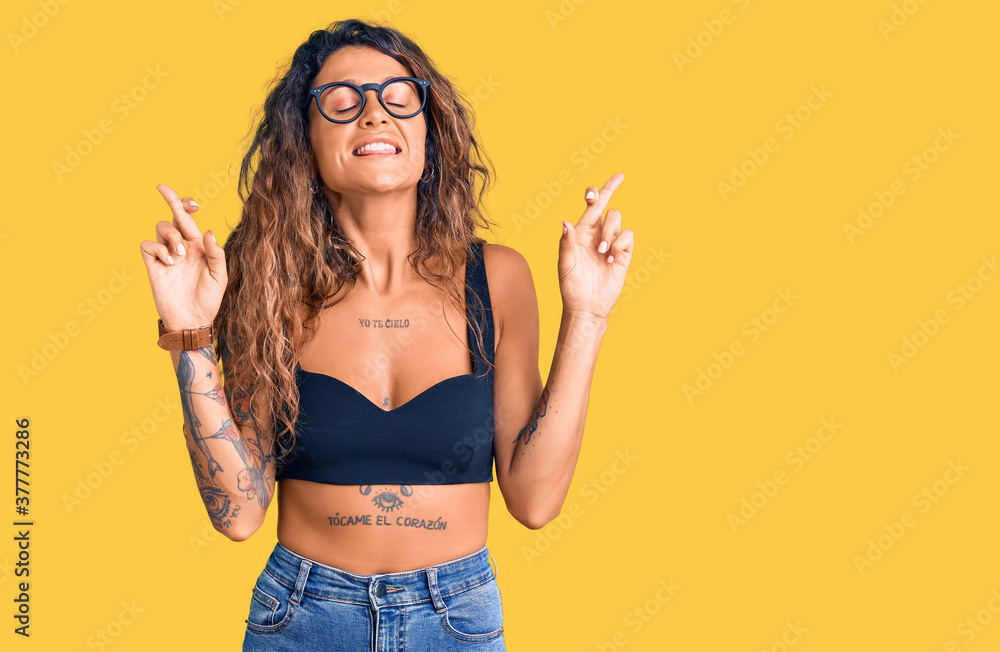 Young hispanic woman with tattoo wearing casual clothes and glasses gesturing finger crossed smiling with hope and eyes closed. luck and superstitious concept. Young hispanic woman with tattoo wearing casual clothes and glasses gesturing finger crossed smiling with hope and eyes closed. luck and superstitious concept.