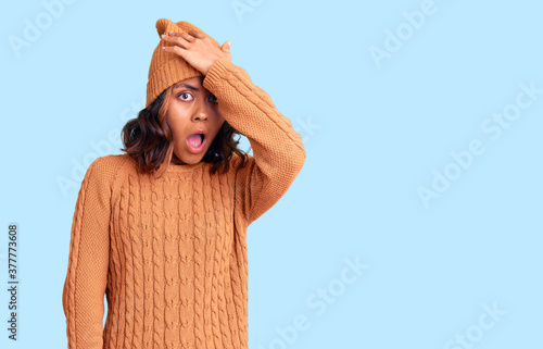 Young beautiful mixed race woman wearing wool sweater and winter hat surprised with hand on head for mistake, remember error. forgot, bad memory concept. photo