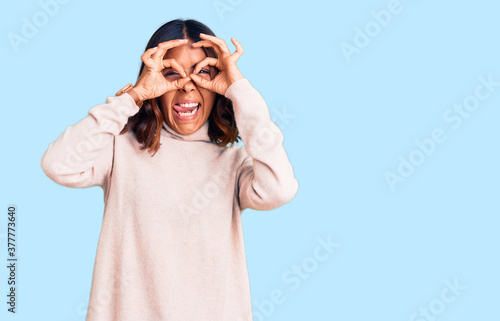 Young beautiful mixed race woman wearing winter turtleneck sweater doing ok gesture like binoculars sticking tongue out, eyes looking through fingers. crazy expression.