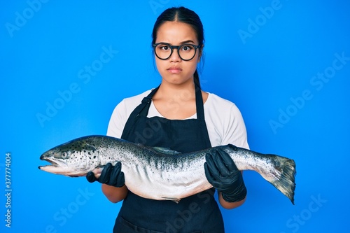 Young beautiful asian girl fishmonger selling fresh raw salmon skeptic and nervous, frowning upset because of problem. negative person.