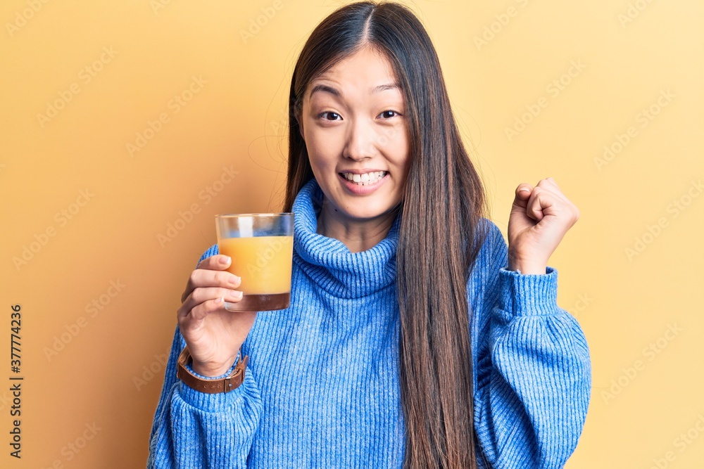 Young beautiful chinese woman drinking glass of orange juice screaming proud, celebrating victory and success very excited with raised arm Young beautiful chinese woman drinking glass of orange juice screaming proud, celebrating victory and success very excited with raised arm