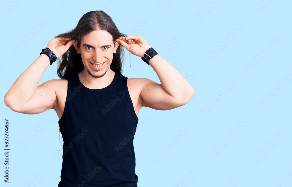 Young adult man with long hair wearing goth style with black clothes smiling pulling ears with fingers, funny gesture. audition problem Young adult man with long hair wearing goth style with black clothes smiling pulling ears with fingers, funny gesture. audition problem