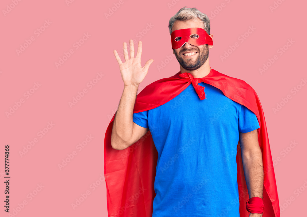 Young blond man wearing super hero custome showing and pointing up with fingers number five while smiling confident and happy. Young blond man wearing super hero custome showing and pointing up with fingers number five while smiling confident and happy.