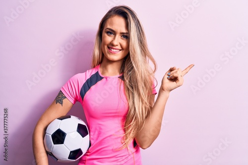 Young beautiful blonde player woman playing soccer holding football ball over pink background smiling happy pointing with hand and finger to the side
