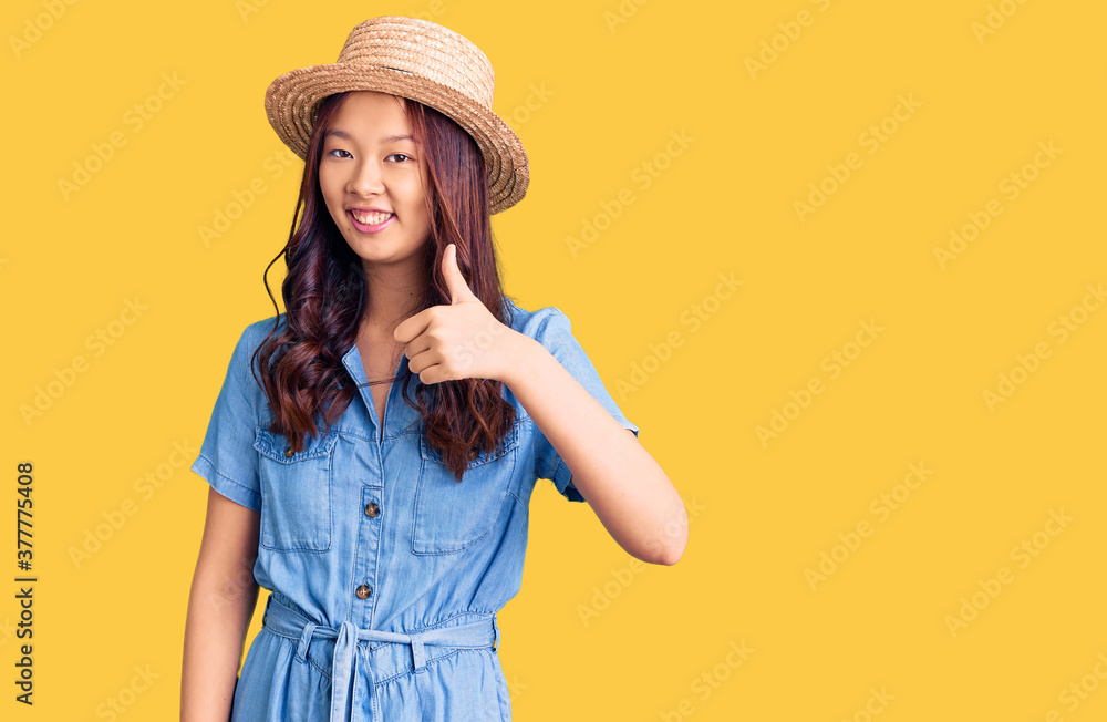Young beautiful chinese girl wearing summer hat doing happy thumbs up gesture with hand. approving expression looking at the camera showing success. Young beautiful chinese girl wearing summer hat doing happy thumbs up gesture with hand. approving expression looking at the camera showing success.