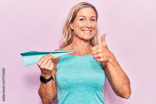 Middle age caucasian blonde woman holding paper airplane smiling happy and positive  thumb up doing excellent and approval sign