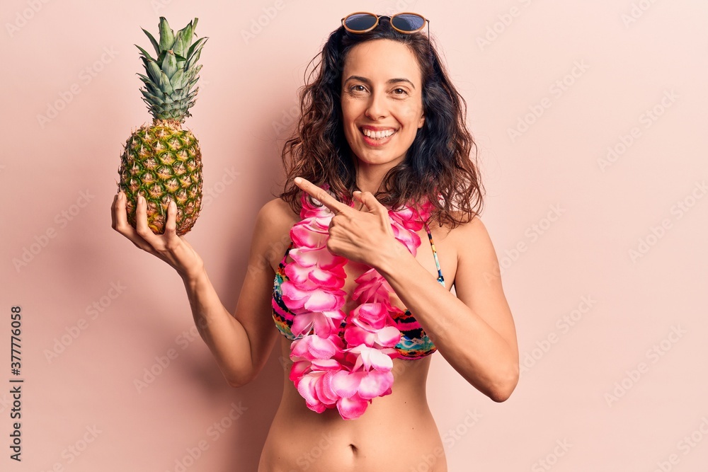 Young beautiful hispanic woman wearing bikini holding pineapple smiling happy pointing with hand and finger Young beautiful hispanic woman wearing bikini holding pineapple smiling happy pointing with hand and finger