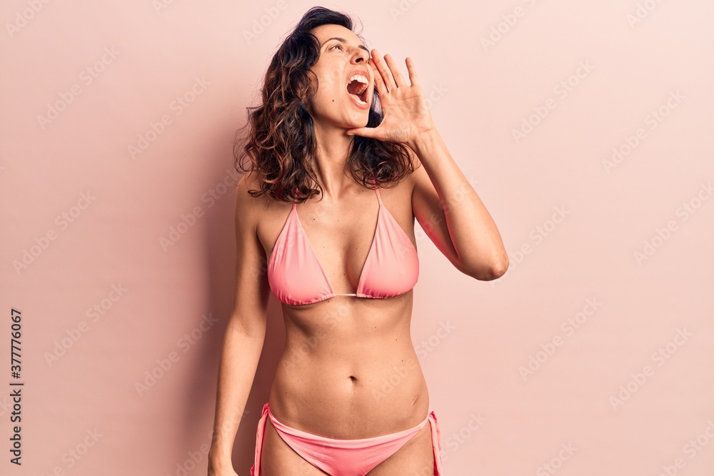 Young beautiful hispanic woman wearing bikini shouting and screaming loud to side with hand on mouth. communication concept. Young beautiful hispanic woman wearing bikini shouting and screaming loud to side with hand on mouth. communication concept.