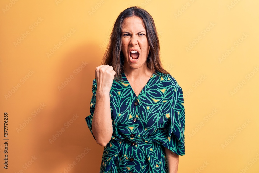 Young beautiful brunette woman wearing casual floral dress standing over yellow background angry and mad raising fist frustrated and furious while shouting with anger. Rage and aggressive concept. Young beautiful brunette woman wearing casual floral dress standing over yellow background angry and mad raising fist frustrated and furious while shouting with anger. Rage and aggressive concept.