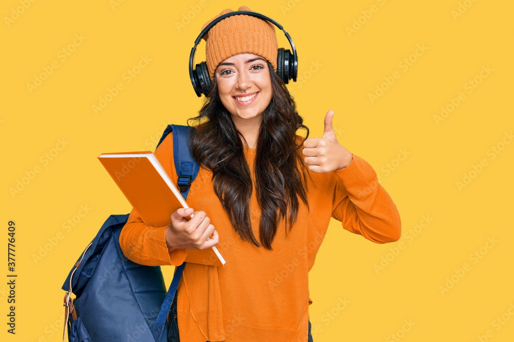Beautiful brunette young woman wearing student backpack and holding books smiling happy and positive, thumb up doing excellent and approval sign Beautiful brunette young woman wearing student backpack and holding books smiling happy and positive, thumb up doing excellent and approval sign