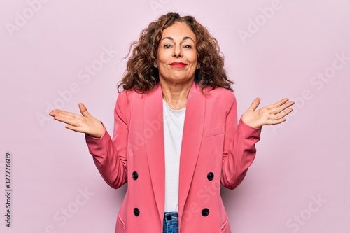 Middle age beautiful businesswoman wearing jacket standing over isolated pink background clueless and confused with open arms, no idea and doubtful face. photo