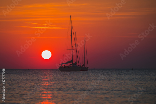 Bloody red sun setting down behind the silohouettes of a two sailboats or yachts moored on an open sea. Adventourous sunset.