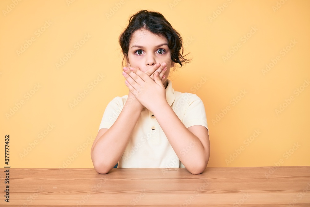 Cute hispanic child wearing casual clothes sitting on the table shocked covering mouth with hands for mistake. secret concept. Cute hispanic child wearing casual clothes sitting on the table shocked covering mouth with hands for mistake. secret concept.