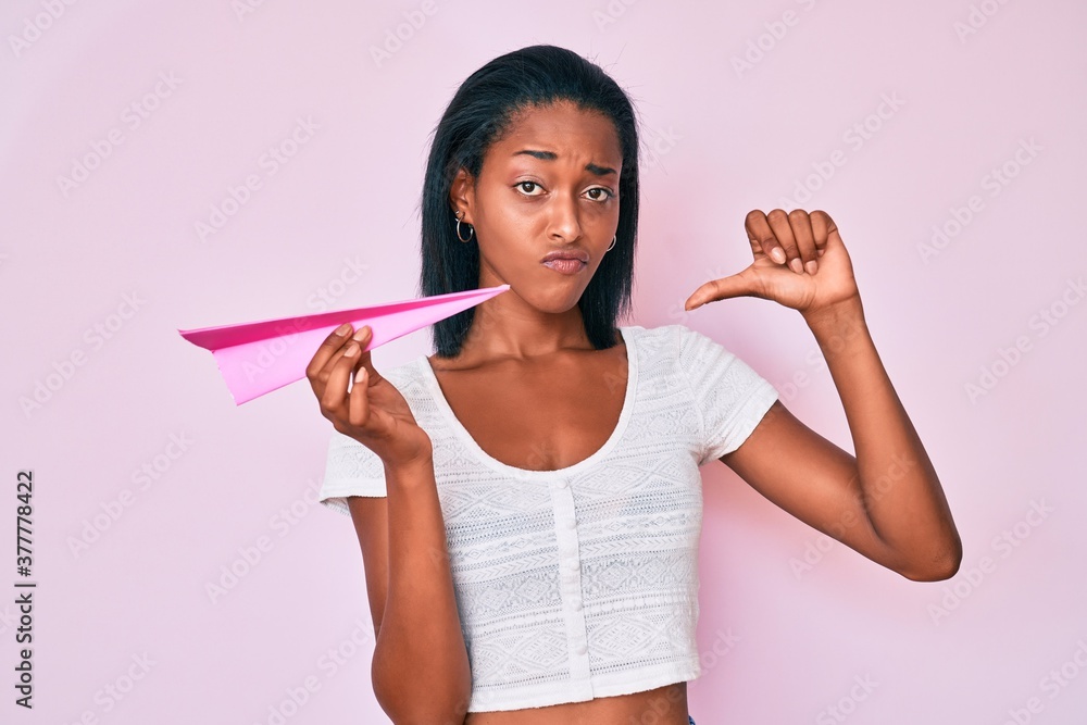 Young african american woman holding paper airplane with angry face, negative sign showing dislike with thumbs down, rejection concept Young african american woman holding paper airplane with angry face, negative sign showing dislike with thumbs down, rejection concept