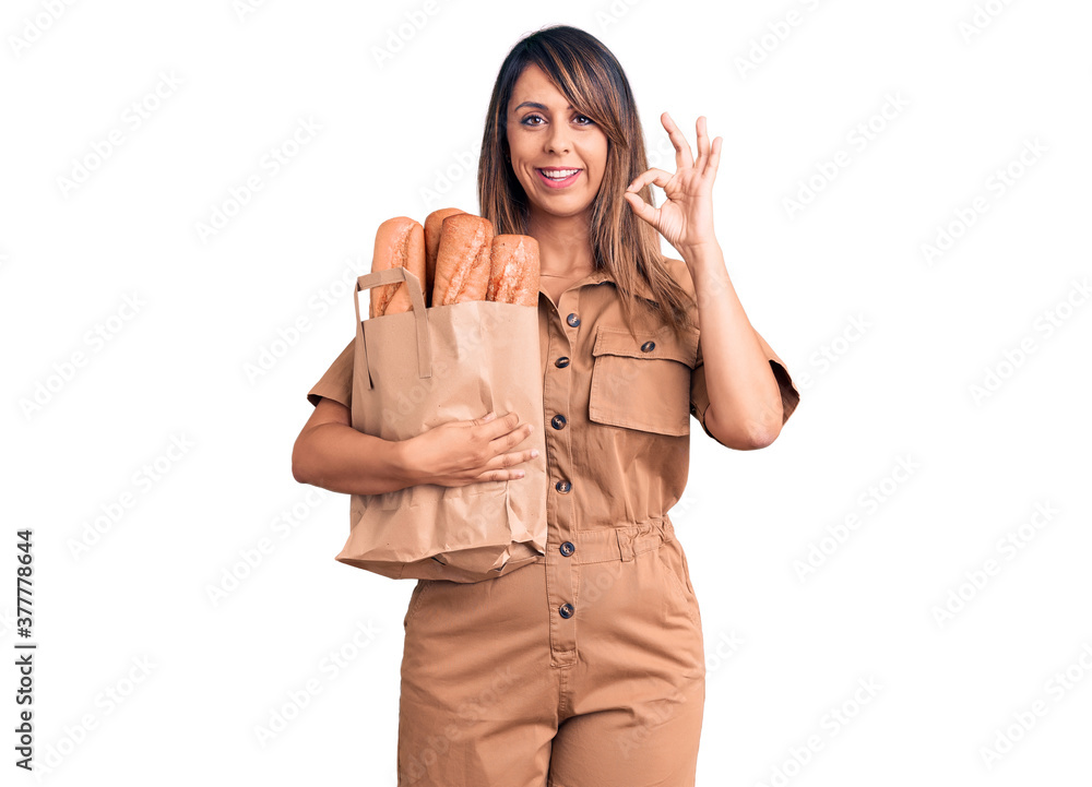 Young beautiful woman holding paper bag with bread doing ok sign with fingers, smiling friendly gesturing excellent symbol Young beautiful woman holding paper bag with bread doing ok sign with fingers, smiling friendly gesturing excellent symbol