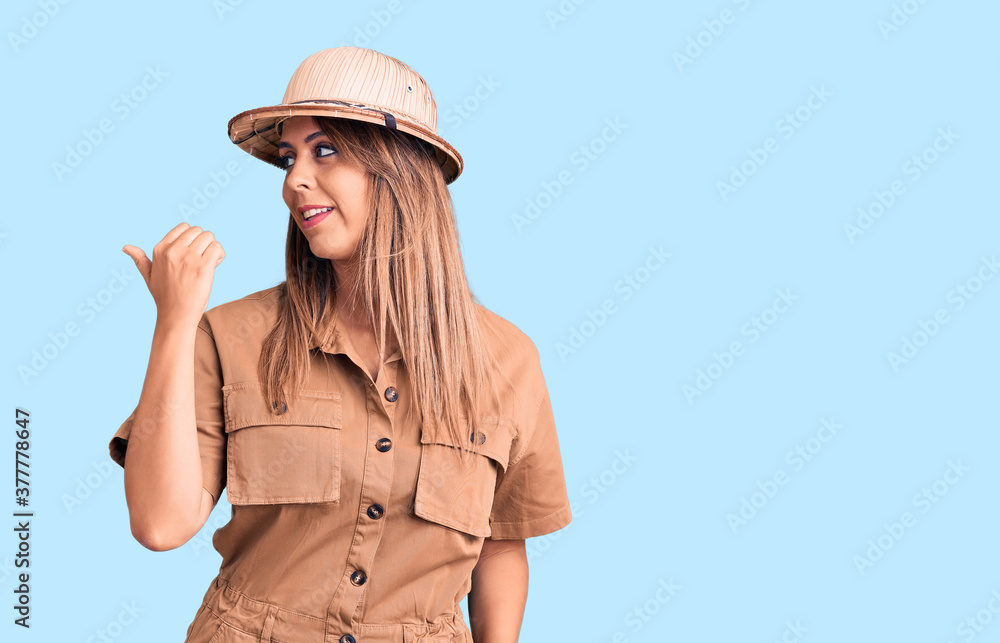 Young beautiful woman wearing explorer hat smiling with happy face looking and pointing to the side with thumb up. Young beautiful woman wearing explorer hat smiling with happy face looking and pointing to the side with thumb up.