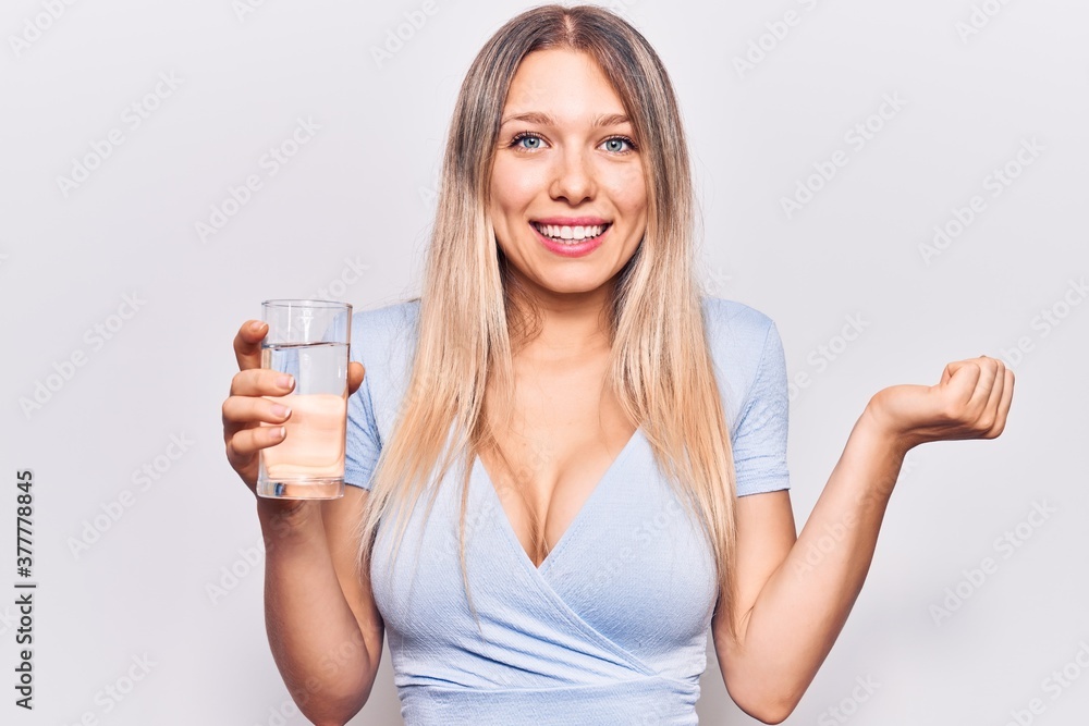 Young beautiful blonde girl drinking glass of water screaming proud, celebrating victory and success very excited with raised arm Young beautiful blonde girl drinking glass of water screaming proud, celebrating victory and success very excited with raised arm