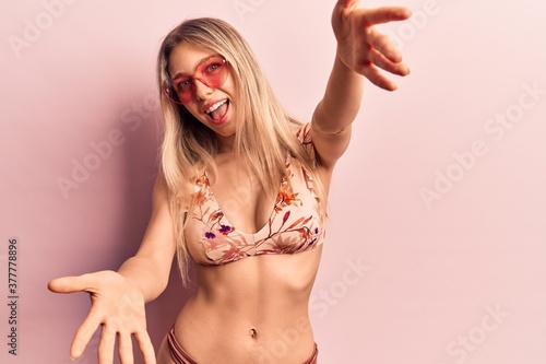 Young beautiful blonde woman wearing bikini heart sunglasses looking at the camera smiling with open arms for hug. cheerful expression embracing happiness.