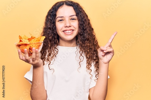 Beautiful kid girl with curly hair holding nachos potato chips smiling happy pointing with hand and finger to the side