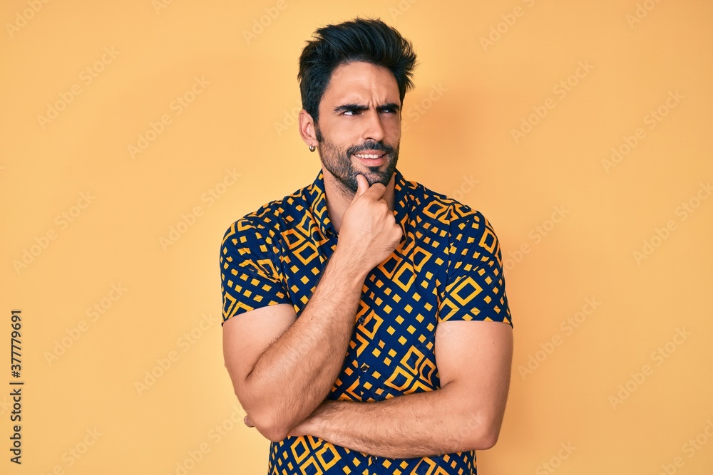 Handsome hispanic man with beard wearing 80s disco style shirt with hand on chin thinking about question, pensive expression. smiling with thoughtful face. doubt concept. Handsome hispanic man with beard wearing 80s disco style shirt with hand on chin thinking about question, pensive expression. smiling with thoughtful face. doubt concept.