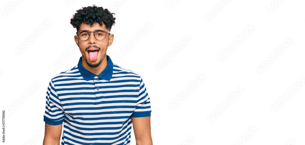 Young arab man wearing casual clothes and glasses sticking tongue out happy with funny expression. emotion concept. Young arab man wearing casual clothes and glasses sticking tongue out happy with funny expression. emotion concept.