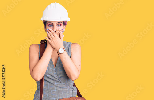 Young beautiful woman wearing architect hardhat and leather bag shocked covering mouth with hands for mistake. secret concept.