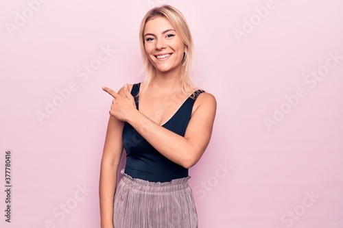 Young beautiful blonde woman wearing casual black t-shirt standing over pink background smiling cheerful pointing with hand and finger up to the side © Krakenimages.com