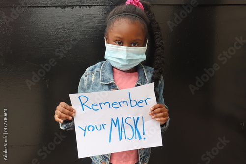 Little black kid wearing face masks holding homemade sign word saying Remember Your Mask black background photo