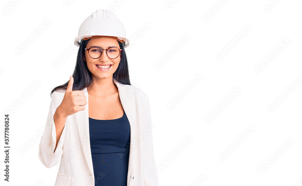 Young beautiful latin girl wearing architect hardhat and glasses doing happy thumbs up gesture with hand. approving expression looking at the camera showing success. Young beautiful latin girl wearing architect hardhat and glasses doing happy thumbs up gesture with hand. approving expression looking at the camera showing success.