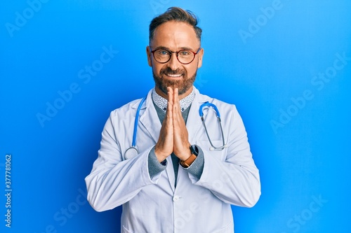 Handsome middle age man wearing doctor uniform and stethoscope praying with hands together asking for forgiveness smiling confident.