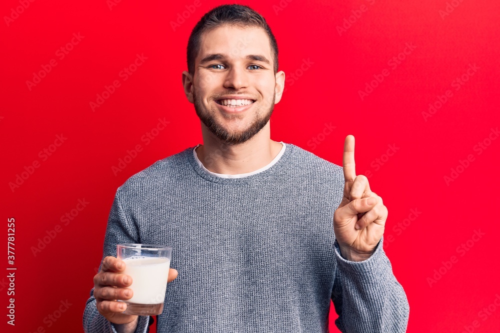 Young handsome man drinking glass of milk smiling with an idea or question pointing finger with happy face, number one Young handsome man drinking glass of milk smiling with an idea or question pointing finger with happy face, number one