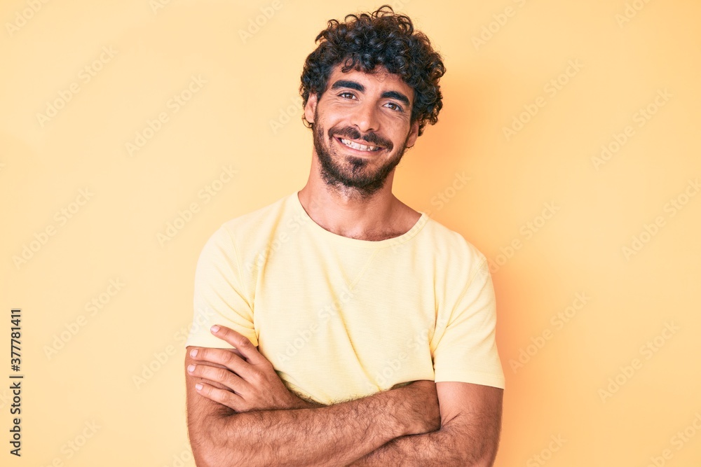 Handsome young man with curly hair and bear wearing casual tshirt over yellow background happy face smiling with crossed arms looking at the camera. positive person. Handsome young man with curly hair and bear wearing casual tshirt over yellow background happy face smiling with crossed arms looking at the camera. positive person.