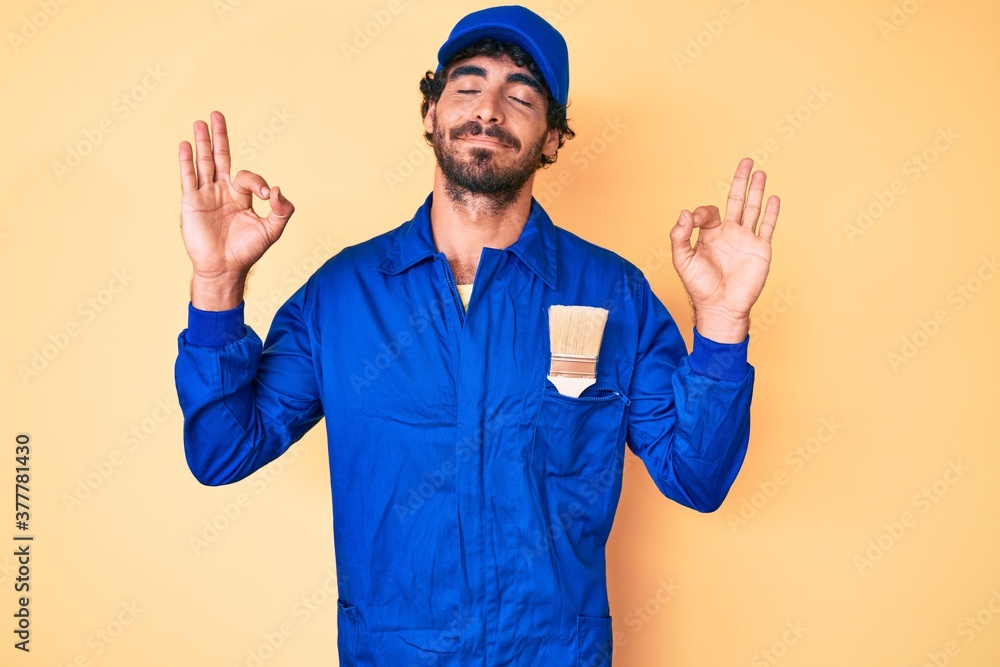 Naklejka premium Handsome young man with curly hair and bear wearing builder jumpsuit uniform relax and smiling with eyes closed doing meditation gesture with fingers. yoga concept.