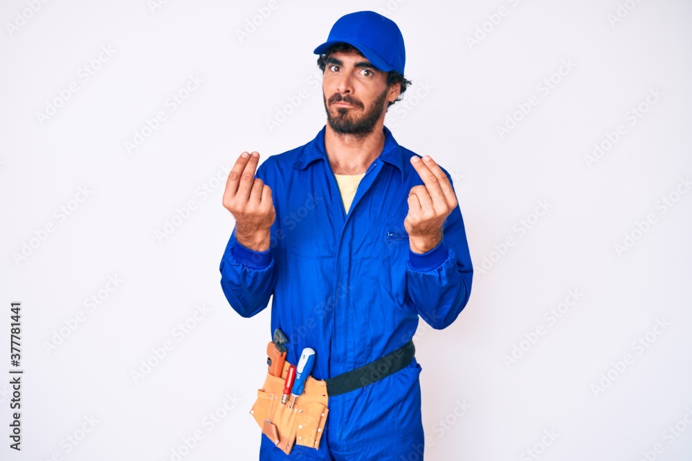 Handsome young man with curly hair and bear weaing handyman uniform doing money gesture with hands, asking for salary payment, millionaire business