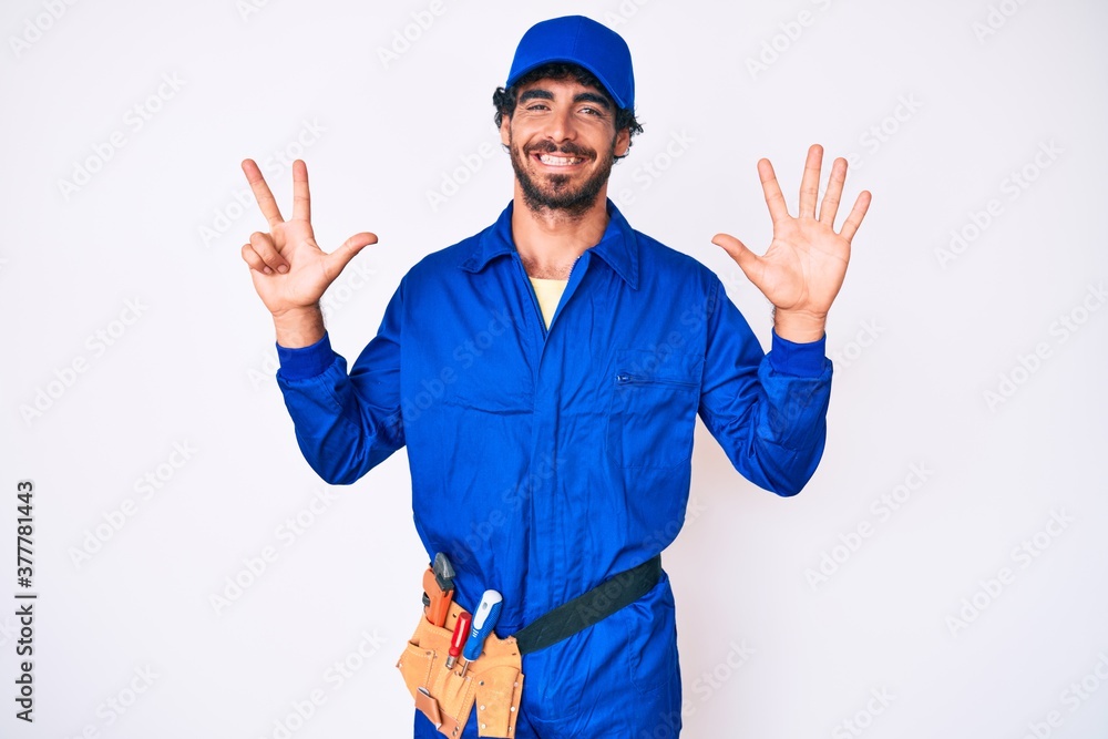 Handsome young man with curly hair and bear weaing handyman uniform showing and pointing up with fingers number eight while smiling confident and happy.