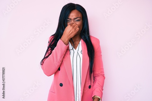 Young african american woman wearing business clothes smelling something stinky and disgusting, intolerable smell, holding breath with fingers on nose. bad smell © Krakenimages.com