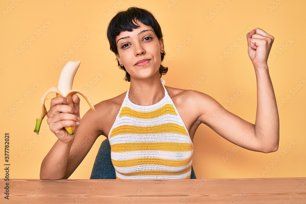 Brunette teenager girl eating banana as healthy snack relaxed with serious expression on face. simple and natural looking at the camera. - obrazy, fototapety, plakaty 