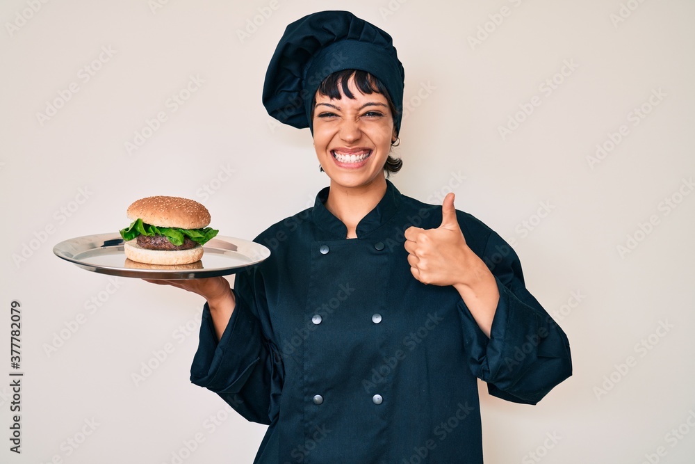 Beautiful brunettte woman chef holding burguer smiling happy and positive, thumb up doing excellent and approval sign Beautiful brunettte woman chef holding burguer smiling happy and positive, thumb up doing excellent and approval sign