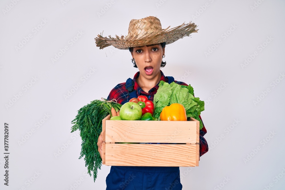 Beautiful brunettte woman wearing farmer clothes holding vegetables in shock face, looking skeptical and sarcastic, surprised with open mouth