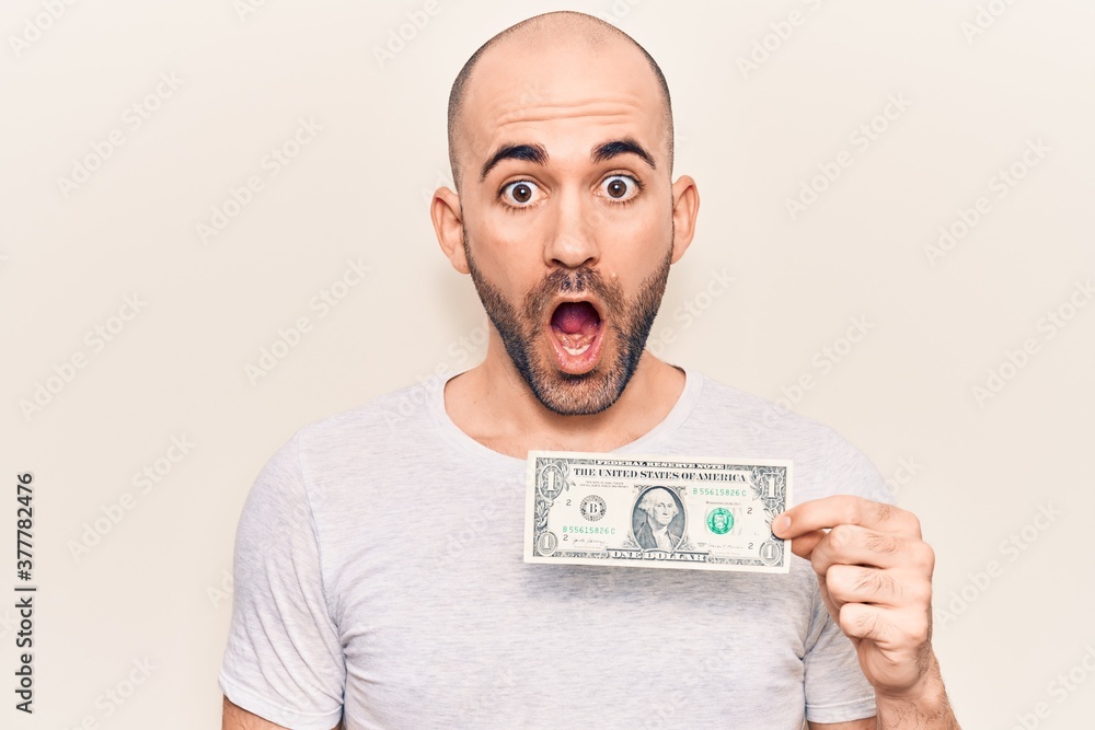 Young handsome bald man holding one dollar banknote scared and amazed with open mouth for surprise, disbelief face Young handsome bald man holding one dollar banknote scared and amazed with open mouth for surprise, disbelief face