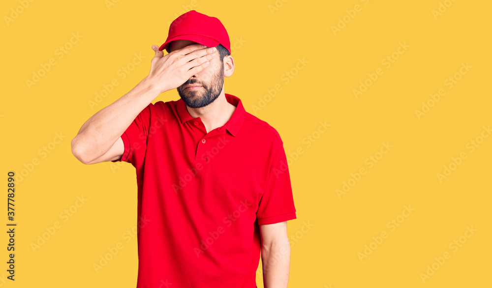 Young handsome man with beard wearing delivery uniform covering eyes with hand, looking serious and sad. sightless, hiding and rejection concept Young handsome man with beard wearing delivery uniform covering eyes with hand, looking serious and sad. sightless, hiding and rejection concept