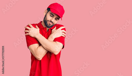 Young handsome man with beard wearing delivery uniform hugging oneself happy and positive, smiling confident. self love and self care © Krakenimages.com