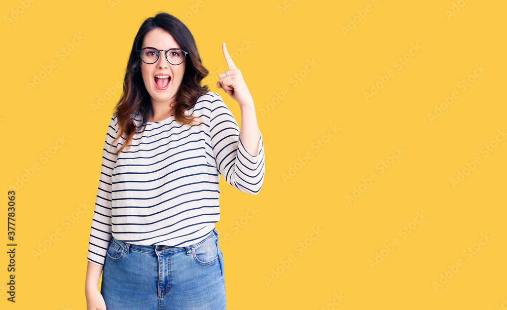 Beautiful young brunette woman wearing casual clothes and glasses pointing finger up with successful idea. exited and happy. number one. Beautiful young brunette woman wearing casual clothes and glasses pointing finger up with successful idea. exited and happy. number one.