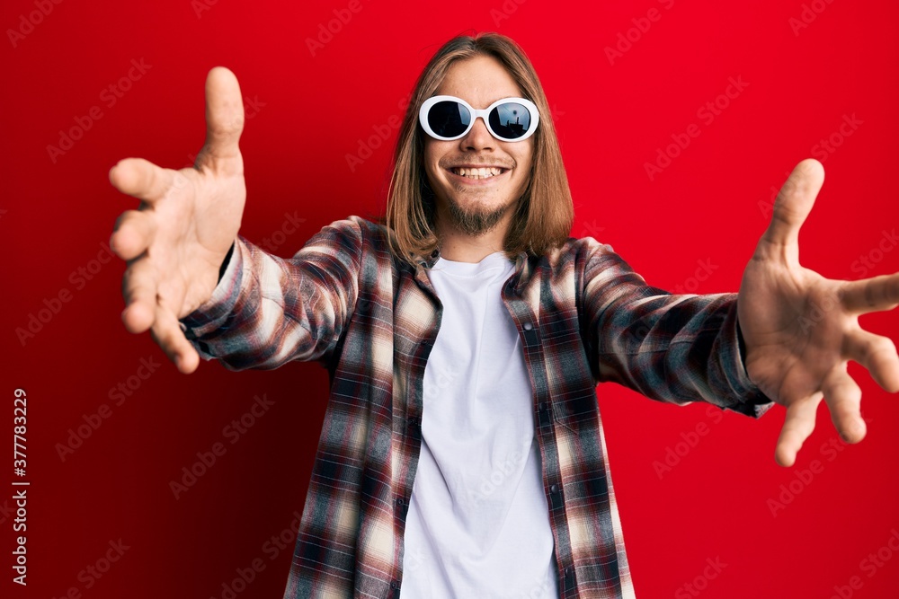 Handsome caucasian man with long hair wearing hipster shirt wearing sunglasses looking at the camera smiling with open arms for hug. cheerful expression embracing happiness. Handsome caucasian man with long hair wearing hipster shirt wearing sunglasses looking at the camera smiling with open arms for hug. cheerful expression embracing happiness.