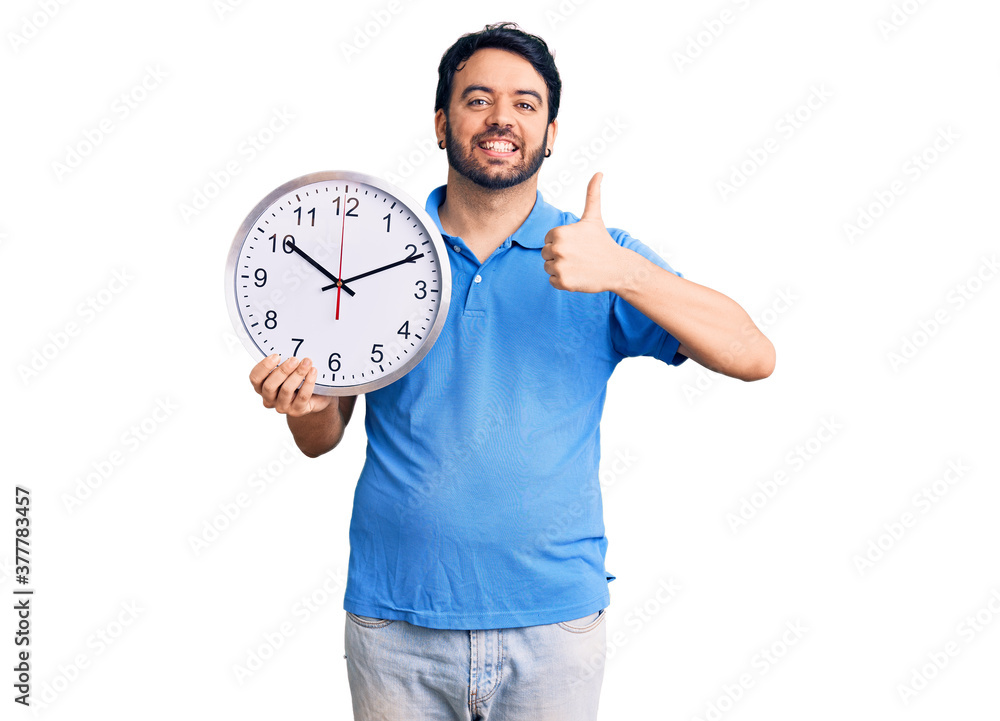 Young hispanic man holding big clock smiling happy and positive, thumb up doing excellent and approval sign Young hispanic man holding big clock smiling happy and positive, thumb up doing excellent and approval sign