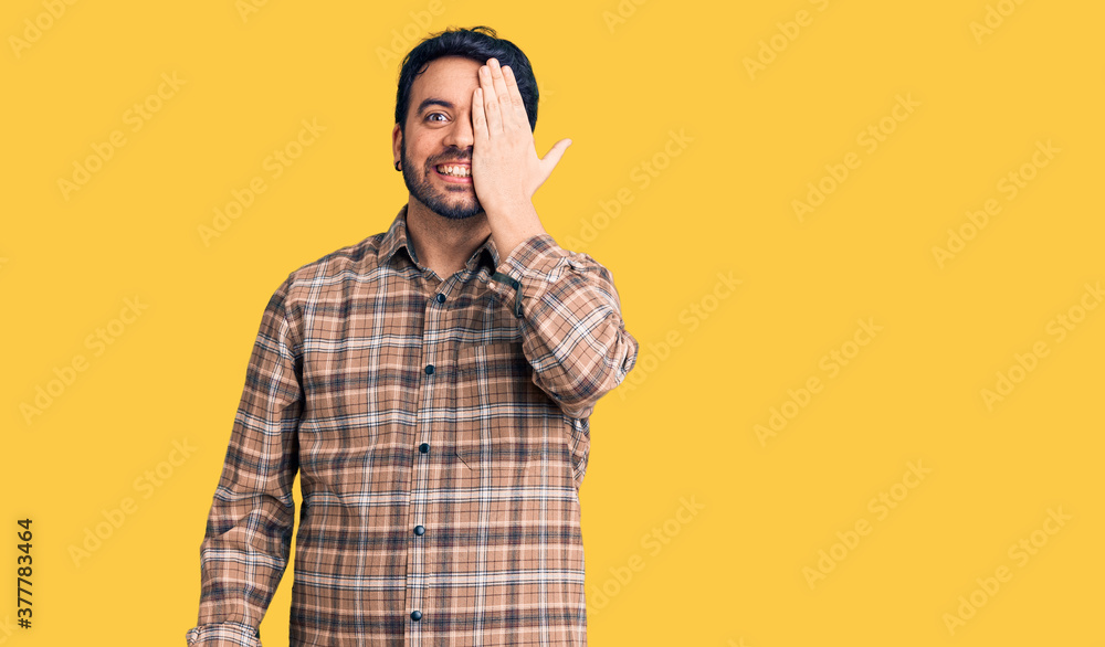 Naklejka premium Young hispanic man wearing casual clothes covering one eye with hand, confident smile on face and surprise emotion.