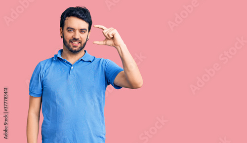 Young hispanic man wearing casual clothes smiling and confident gesturing with hand doing small size sign with fingers looking and the camera. measure concept. © Krakenimages.com