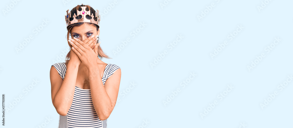 Young beautiful woman wearing king crown shocked covering mouth with hands for mistake. secret concept.